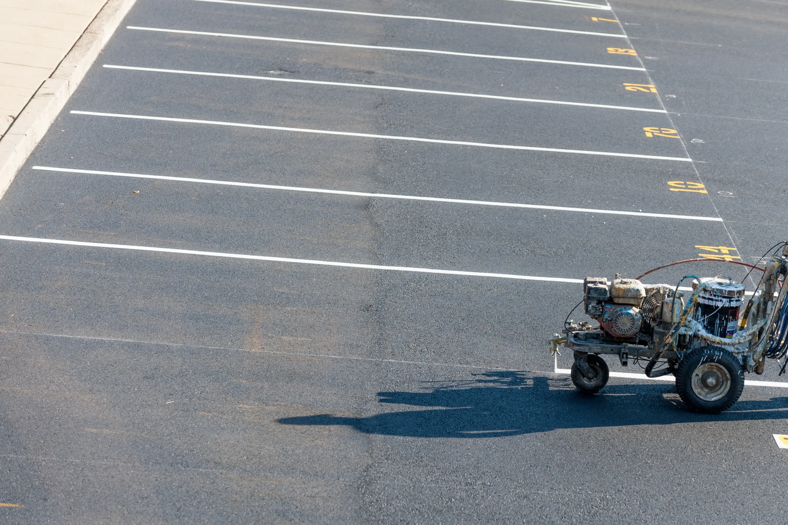 A machine is painting white lines on a freshly paved parking lot. Numbered spaces are visible, with concrete curbs on the left, under a bright, sunny sky.