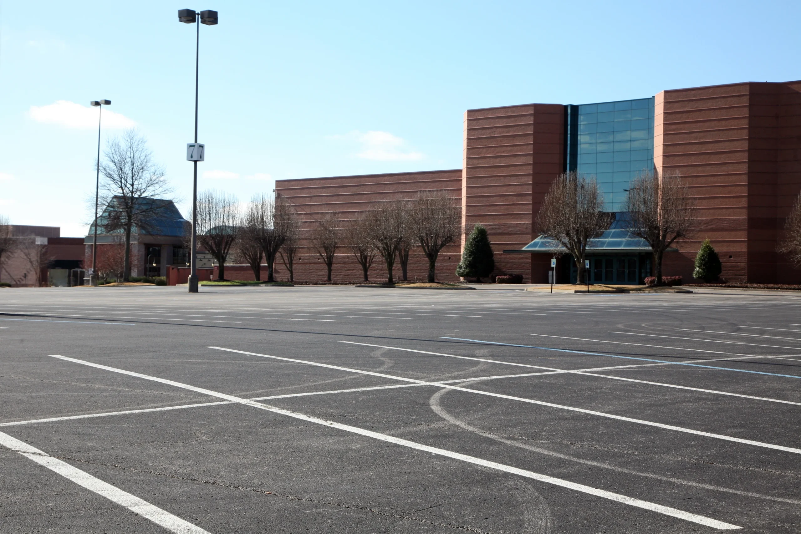 Empty parking lot in front of a large, modern brick building with tall windows. Bare trees and lampposts are visible under a clear blue sky.